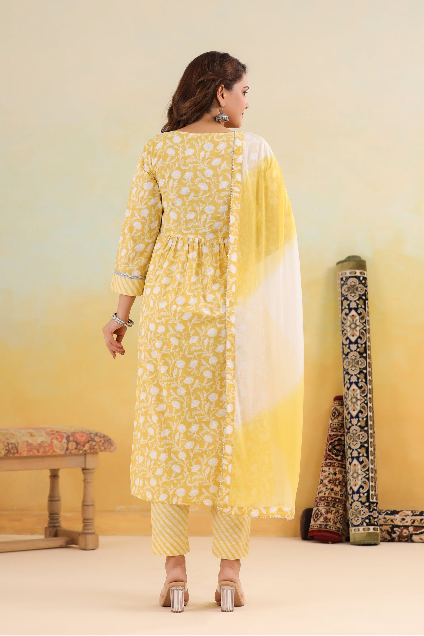 Woman in a yellow floral dress standing in a room with a yellow wall and decorative items.
