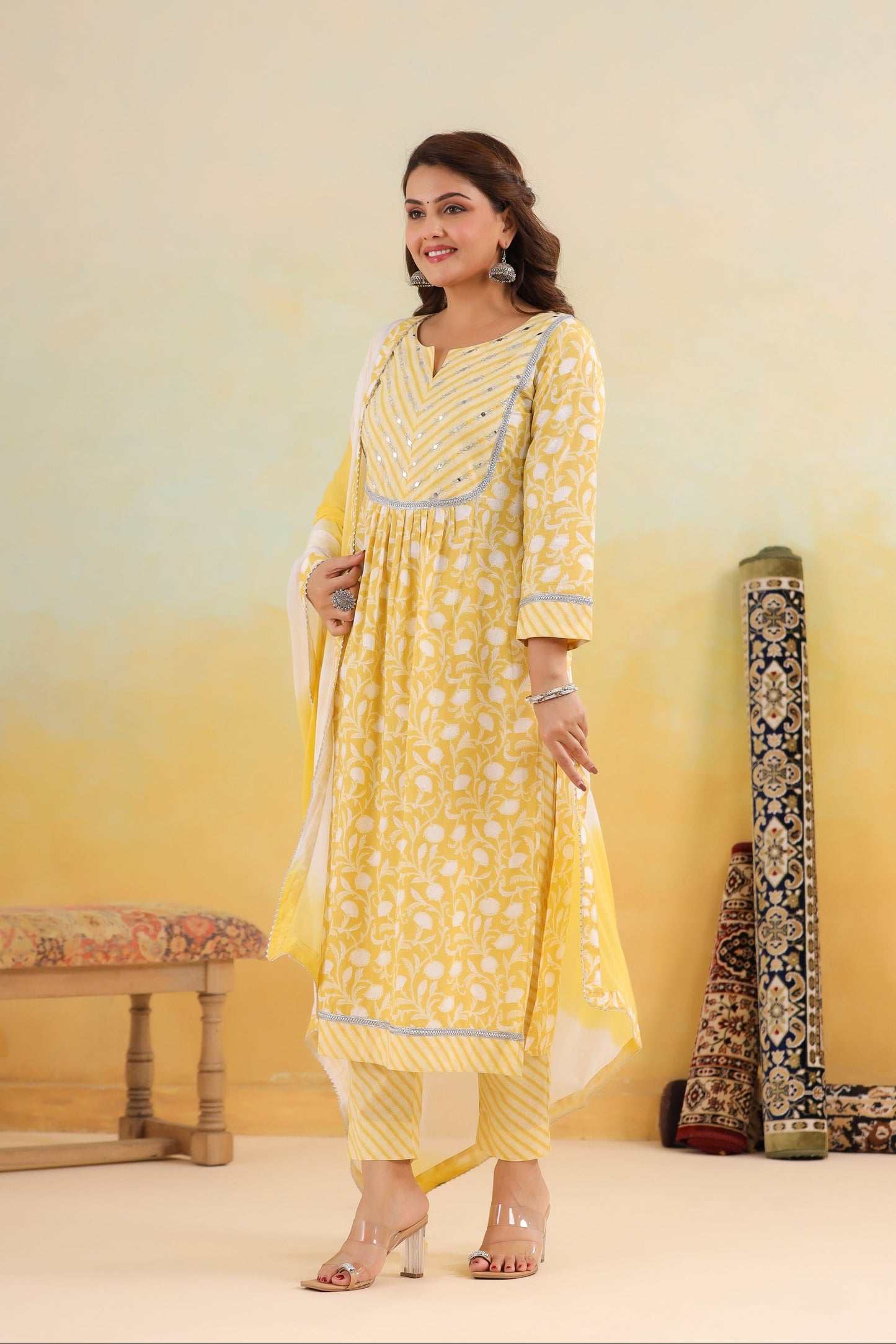 Woman in a yellow traditional outfit standing next to decorative fabrics against a light yellow background