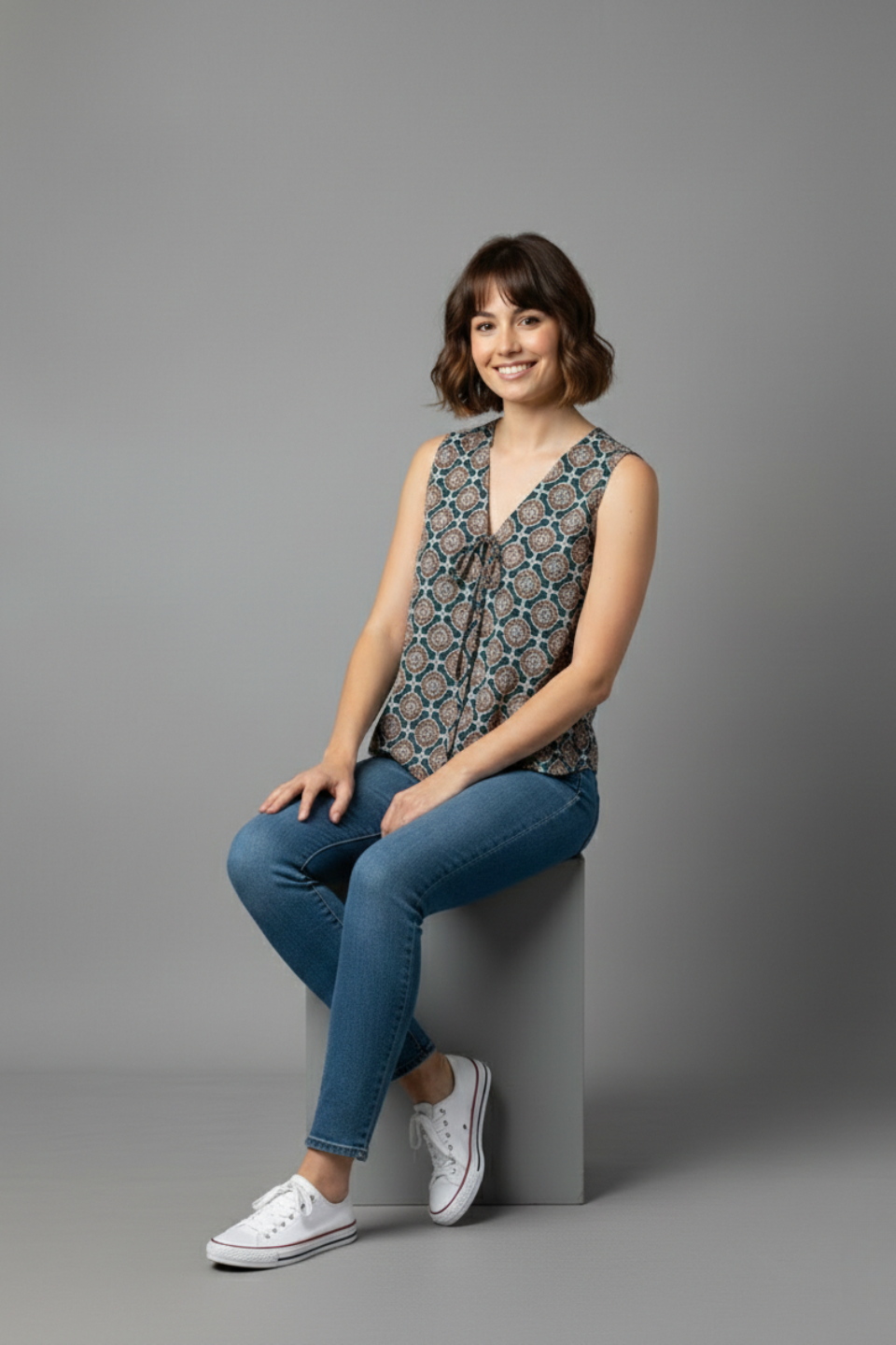 Woman wearing printed sleeveless top, jeans, and white sneakers, seated on gray block