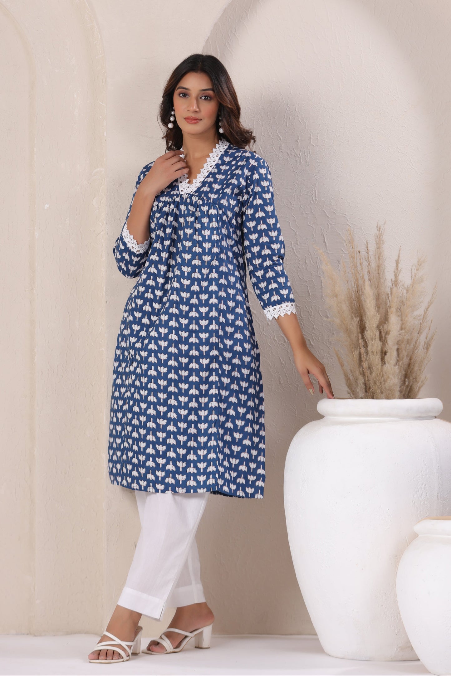 Woman wearing a blue patterned dress standing next to a white vase with dried plants.