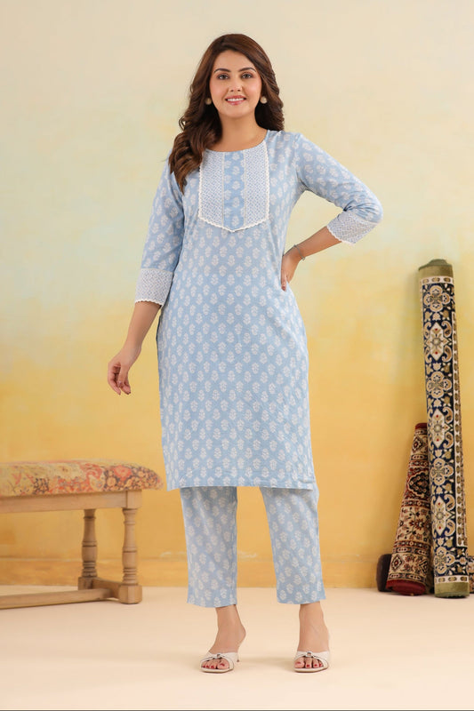 Woman wearing a light blue kurta with white patterns in a room with a wooden bench and decorative items.